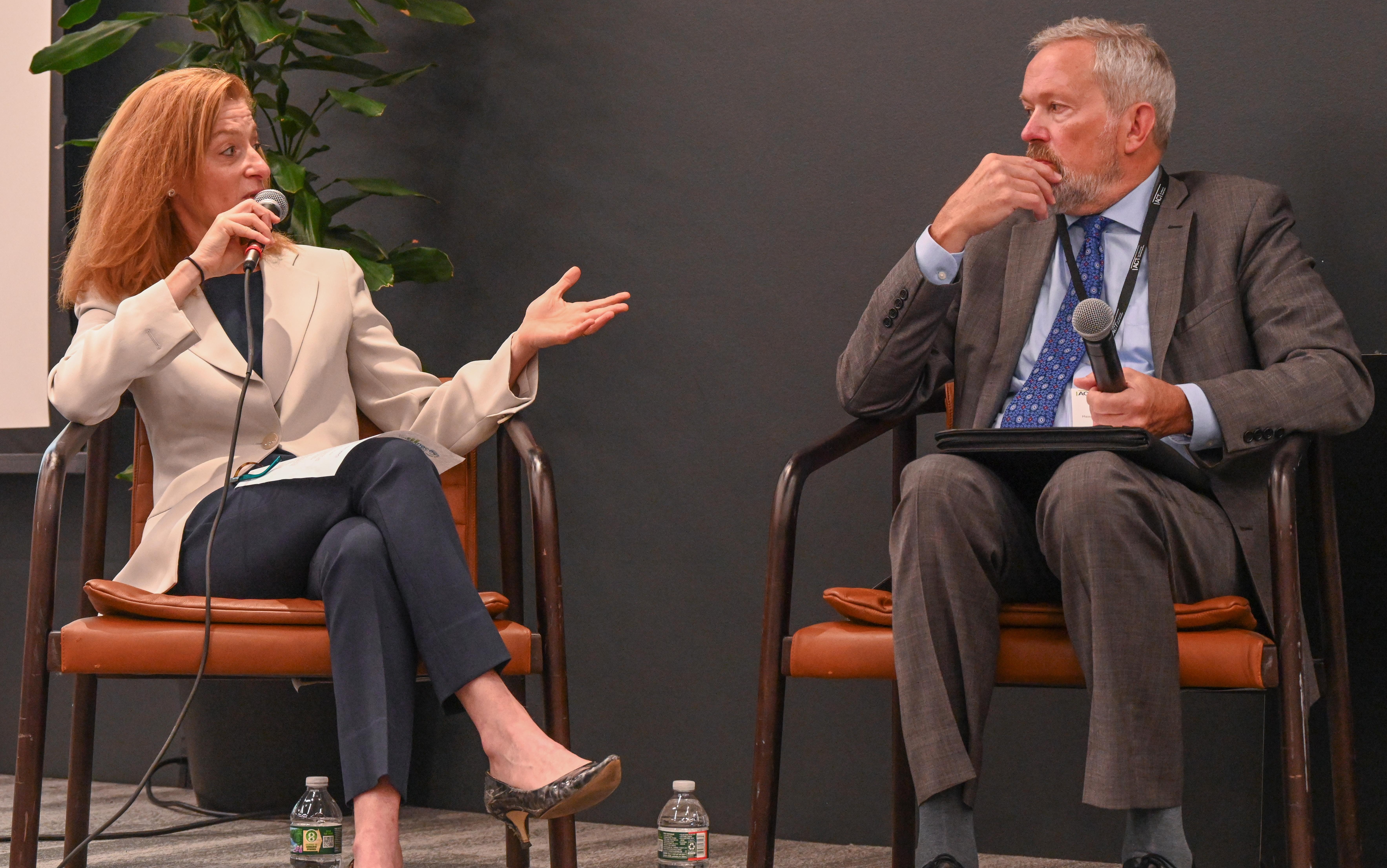 A woman and a man wearing business attire seated in chairs each holding a microphone with woman speaking