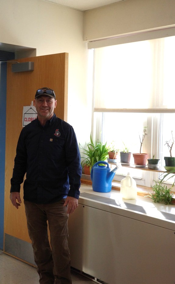 A man stands beside a classroom window with potted plants and watering cans above a ventilation unit.