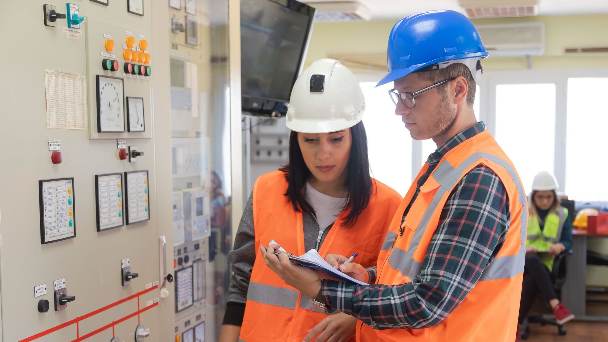 A male and female engineer, both wearing high-visibility vests and safety helmets, collaborate in the bright control room of a renewable energy facility