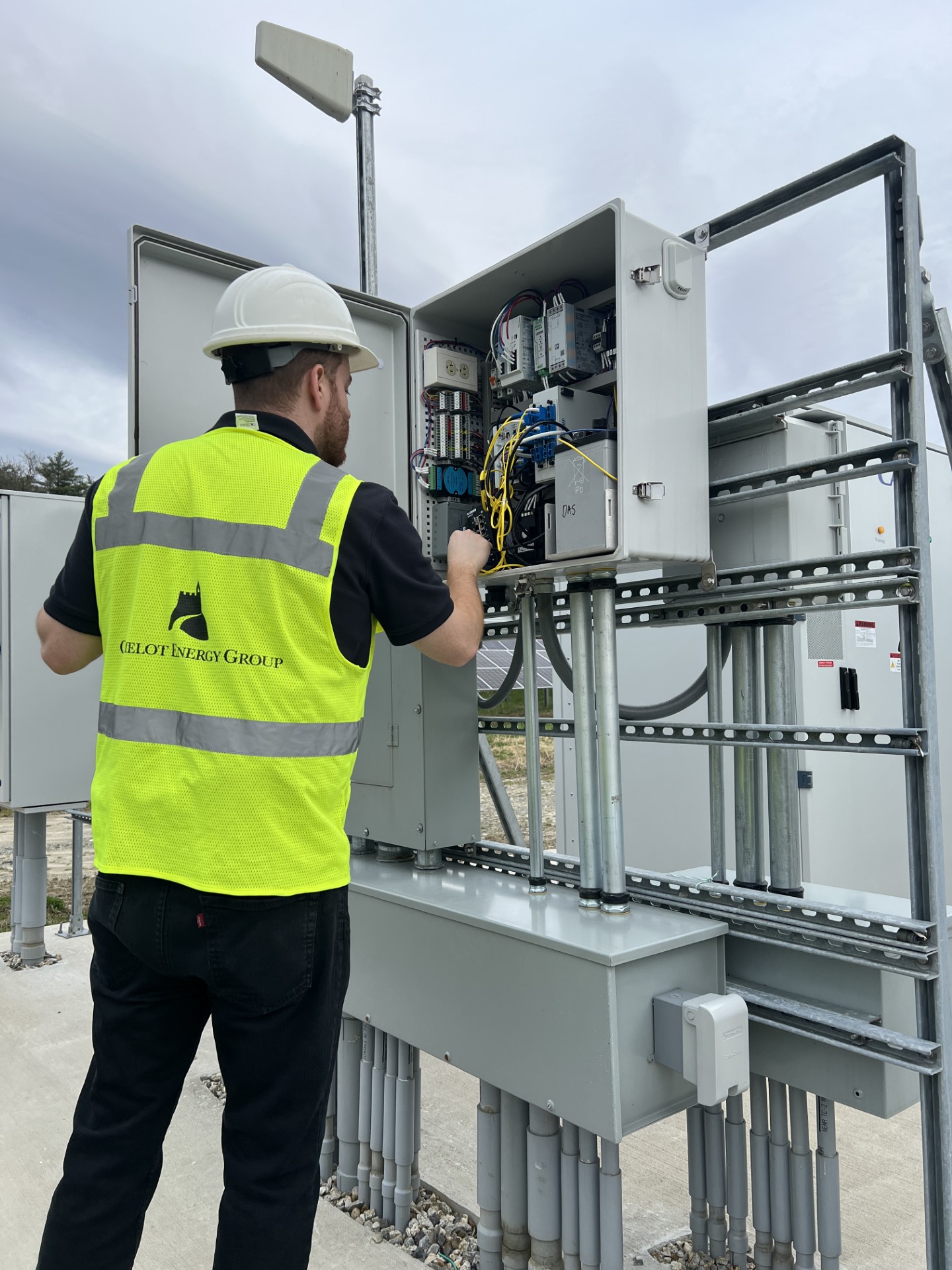 A worker wearing a hard hat and high-visibility vest inspects wiring inside an open electrical control cabinet at an outdoor clean energy installation.