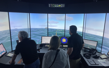 Three people observe an offshore wind vessel simulator showing a virtual ocean scene with wind turbines and navigation controls.