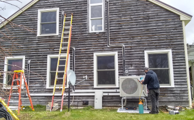 An air-source heat pump outdoor unit being installed outside a brown house