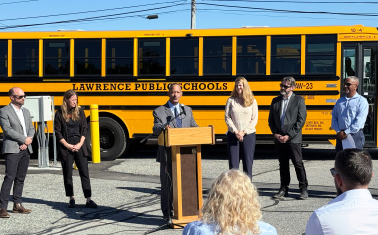People standing in front of a parked electric school bus during an event. Speaker at a podium addresses a crowd.