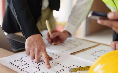 People working on paper architectural plans with computer, calculator and hardhat