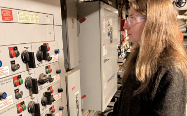 A woman wearing safety glasses reviews equipment inside a mechanical room while standing beside an electrical control panel.