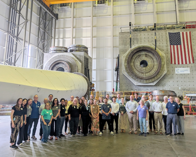 Members of the Offshore Energy Community of Practice visit the Wind Technology Testing Center, standing beside a utility-scale wind turbine blade and structural testing equipment inside the facility.