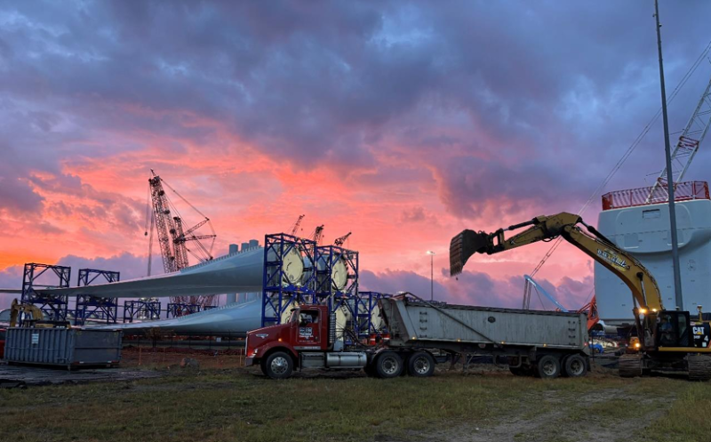 An excavator loads a dump truck, to the left large wind turbine blades lay stacked together