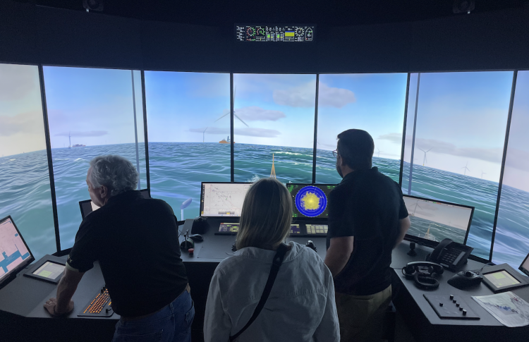 Three people observe an offshore wind vessel simulator showing a virtual ocean scene with wind turbines and navigation controls.