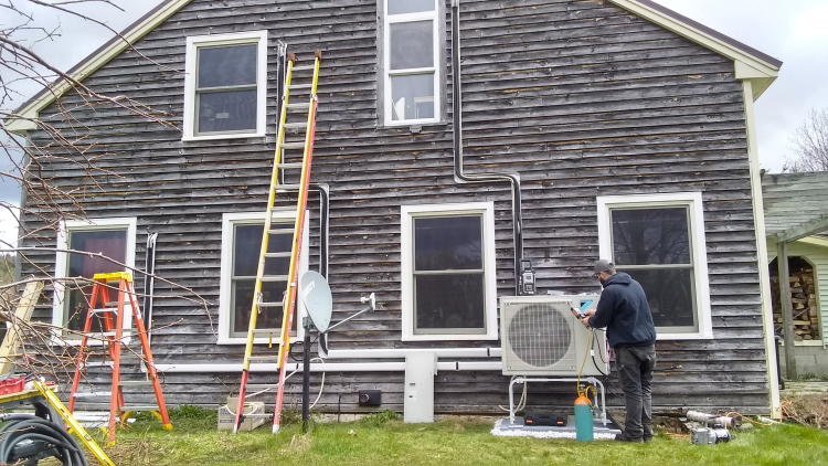 An air-source heat pump outdoor unit being installed outside a brown house