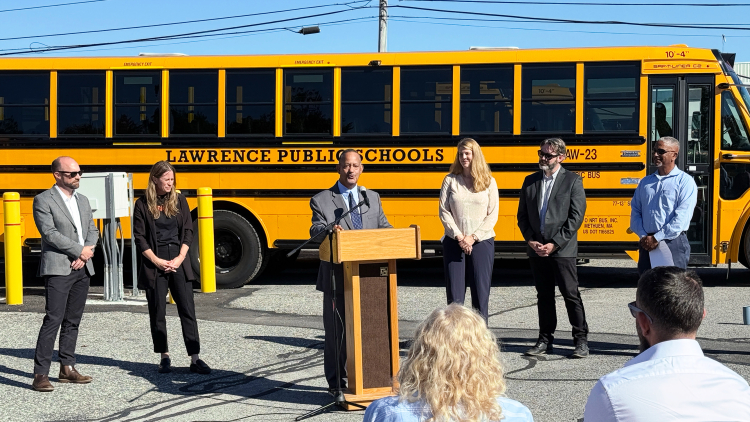 People standing in front of a parked electric school bus during an event. Speaker at a podium addresses a crowd.