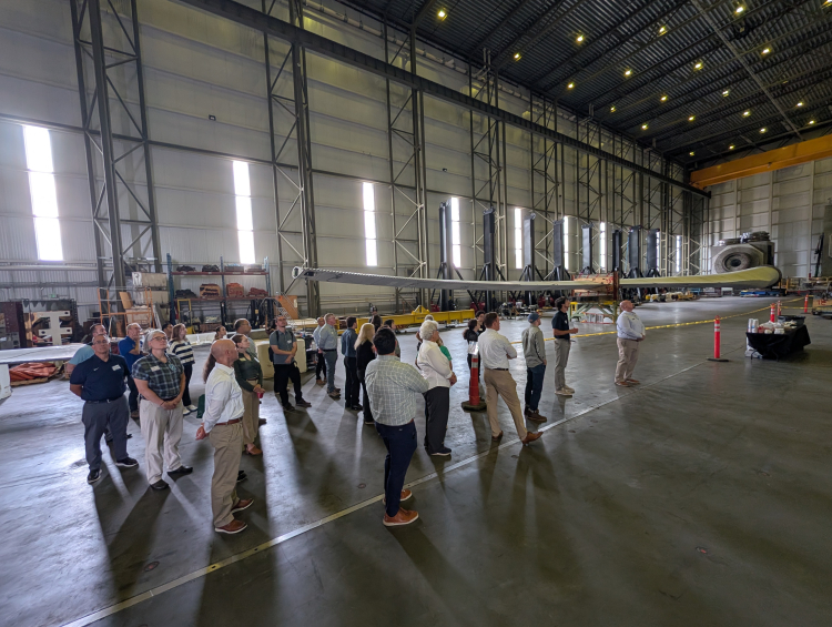 Offshore Wind Workforce Community of Practice members tour a large wind turbine testing facility, standing beneath a turbine blade.