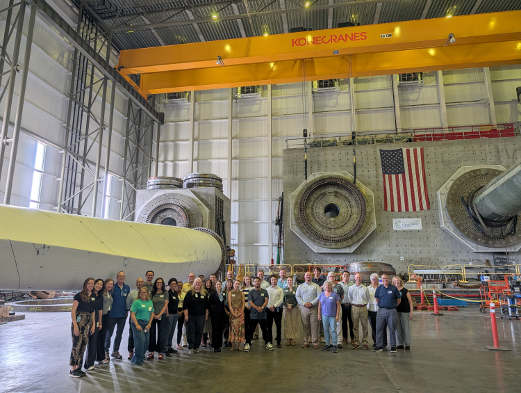 Offshore Wind Workforce Community of Practice members pose for a group photo inside a wind turbine testing facility, with large turbine components behind them.