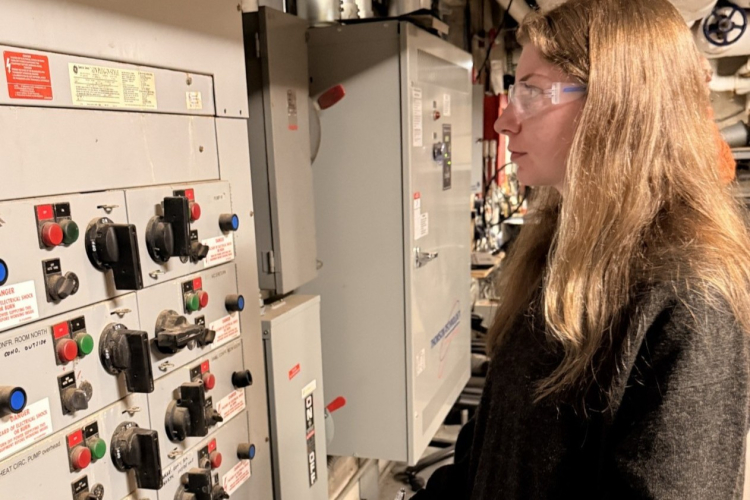 A woman wearing safety glasses reviews equipment inside a mechanical room while standing beside an electrical control panel.