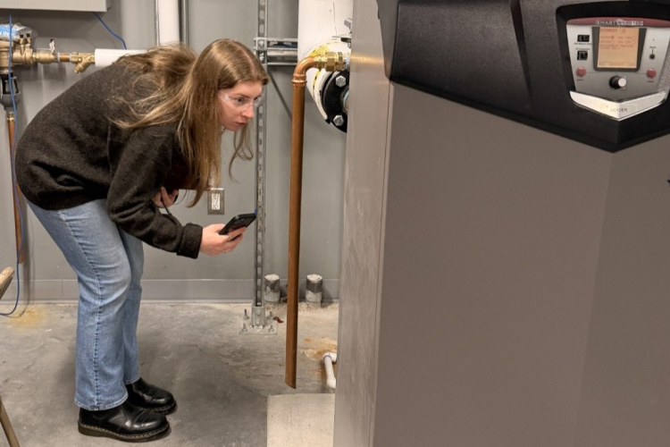 A woman wearing safety glasses examines mechanical equipment in a utility room while holding a phone.