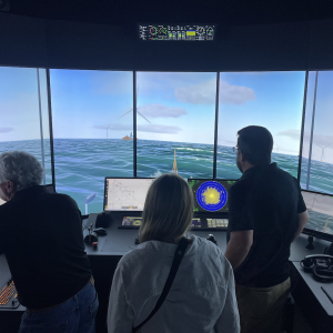 Three people observe an offshore wind vessel simulator showing a virtual ocean scene with wind turbines and navigation controls.