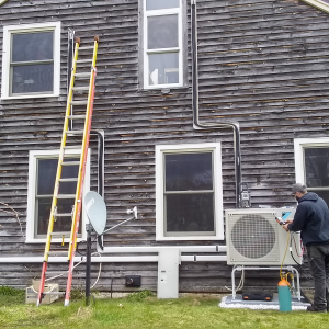 An air-source heat pump outdoor unit being installed outside a brown house