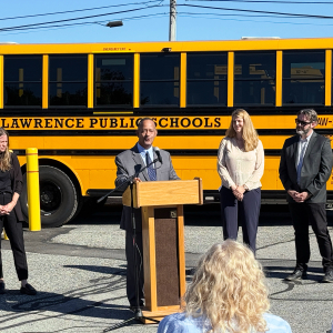 People standing in front of a parked electric school bus during an event. Speaker at a podium addresses a crowd.