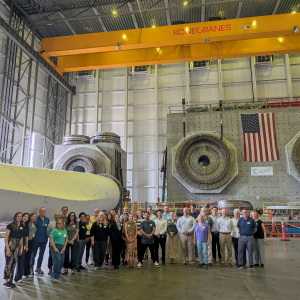 Offshore Wind Workforce Community of Practice members pose for a group photo inside a wind turbine testing facility, with large turbine components behind them.