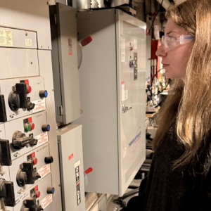 A woman wearing safety glasses reviews equipment inside a mechanical room while standing beside an electrical control panel.