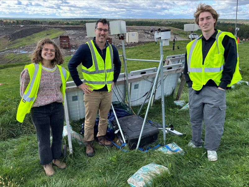 Three people standing with a gas monitoring system in a landfill.