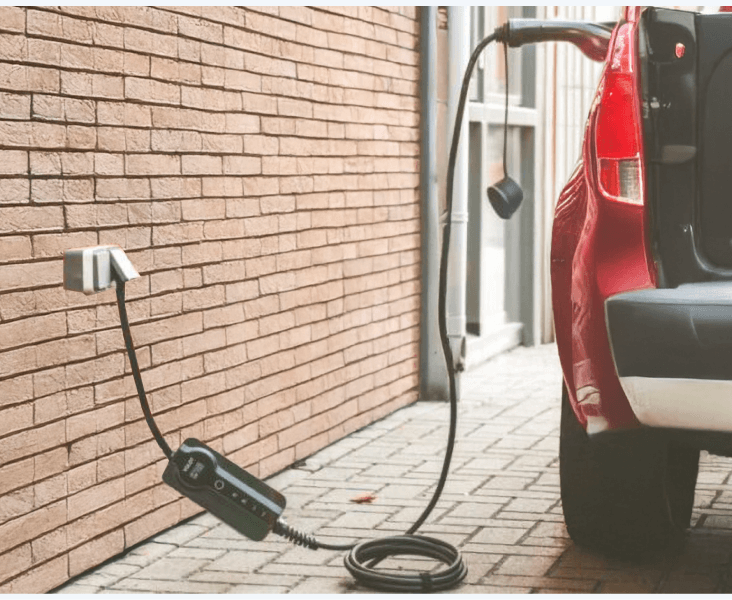 Electric car charging from a wall outlet with portable EV charger cable beside a brick building.