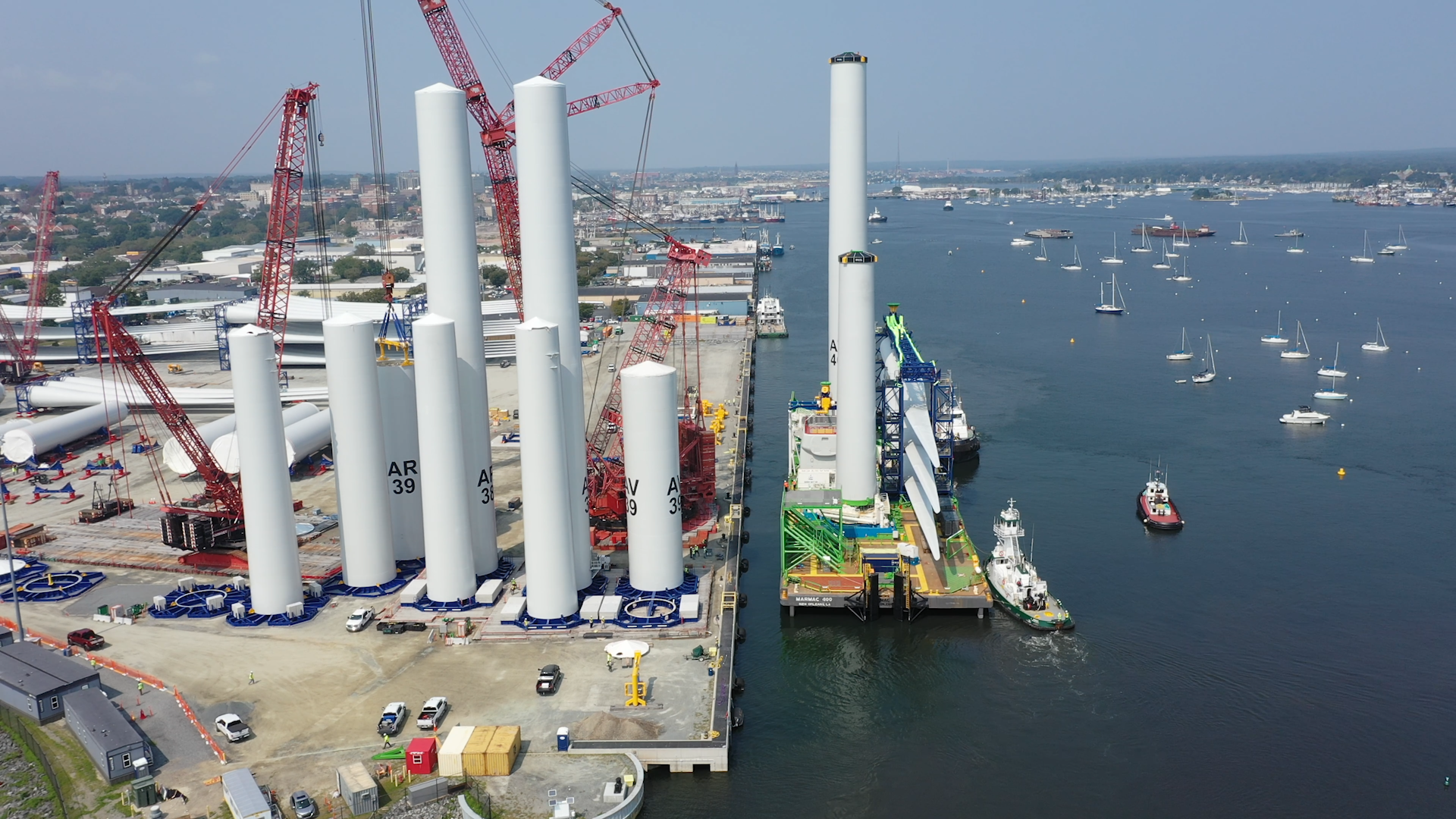 Wind turbine towers standing vertically at New Bedford Marine Commerce Terminal and being moved with tall red cranes