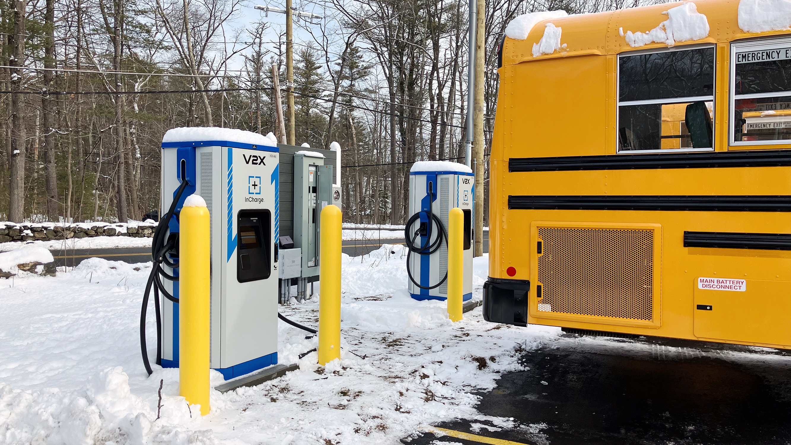 A yellow school bus parked beside three V2X electric vehicle charging stations at a snowy outdoor bus depot.