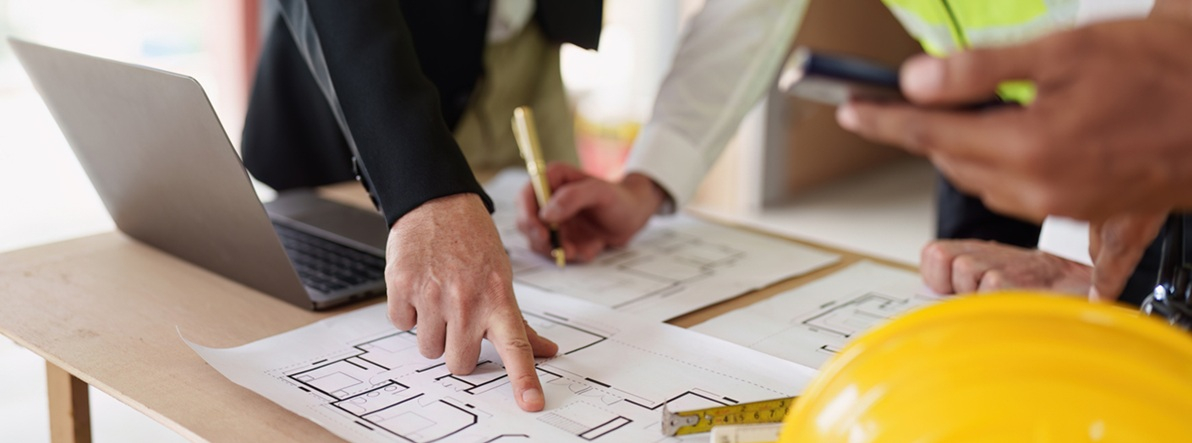 People looking at design drawings on a desk with laptop, ruler and hardhat