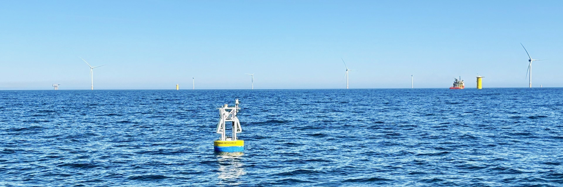 Ocean research buoy in the foreground with offshore wind turbines and support vessels on the horizon, illustrating offshore wind energy development and marine research in coastal waters.