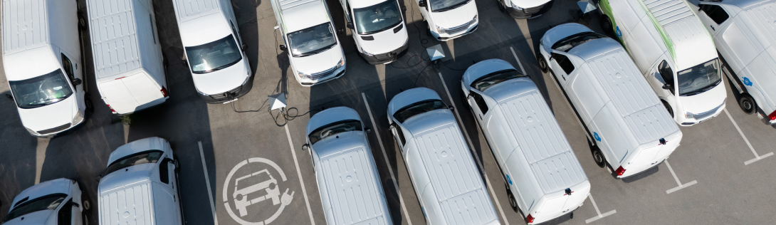 Two rows of commercial electric vehicles are parked in a parking lot, some of which are charging.